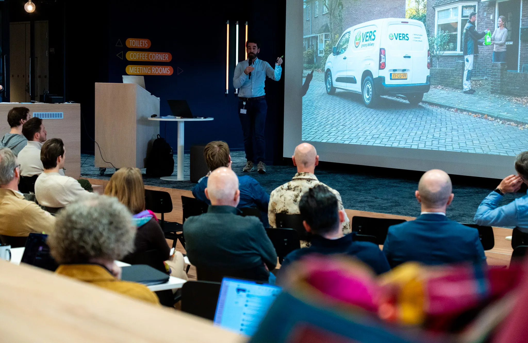 Koen de Greef presenting to a seated audience, gesturing towards a design case on the screen behind him.